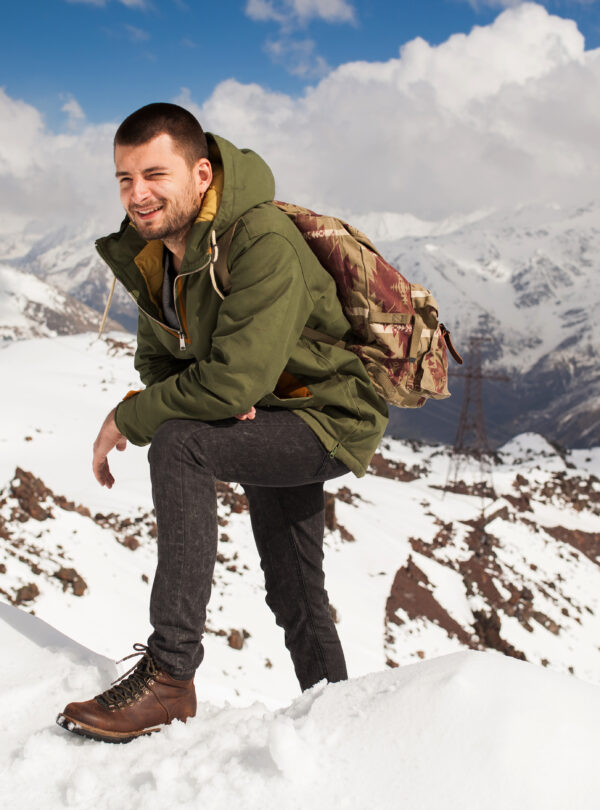 young hipster man hiking in mountains, winter vacation traveling, north, snow, warm clothes, backpack, anorak, jeans, boots
